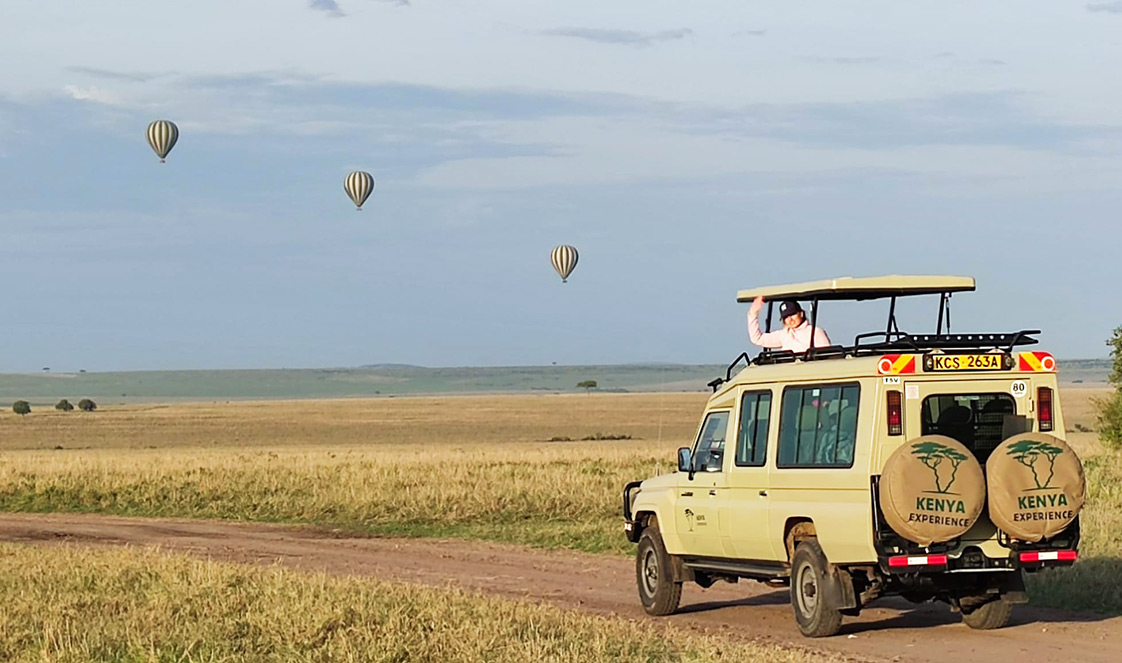 A safari vehicle with a guest waving from the pop up roof, three hot air balloons on the horizon.