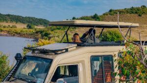 Two people looking at the Mara River through the pop up roof of their safari vehicle.