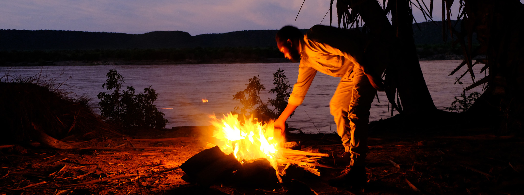 A man lights a campfire near a river in Tsavo East. Budgeting for a Kenya safari is essential in planning your trip.