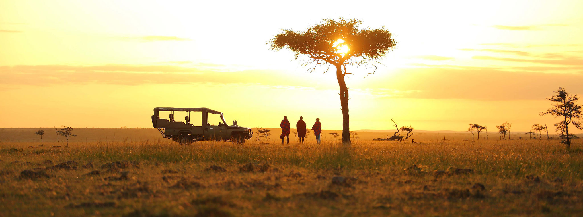 A safari vehicle and guests standing under an acacia tree at sunset. Sundowner drinks are a tradition on Kenya luxury safaris.