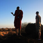 A Maasai guide points at something in the distance while a guest looks on. Both are on a bush walk in the Amboseli region.