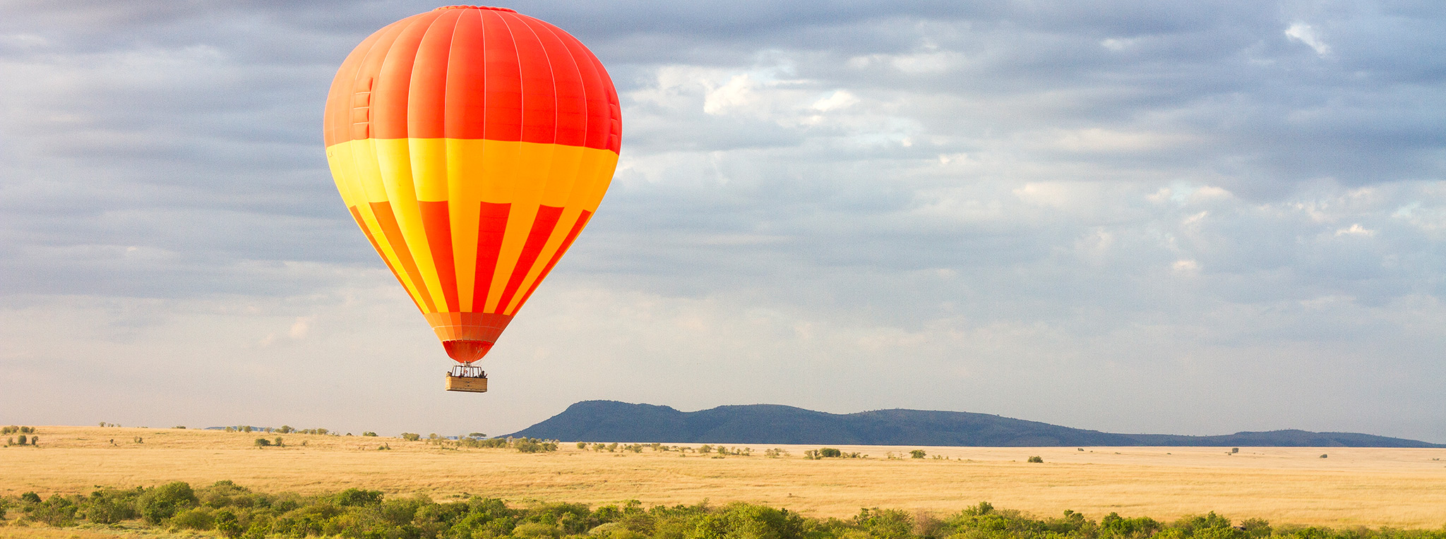 An orange and yellow hot air balloon floats across the savanna of the Maasai Mara on a hot air balloon safari in Kenya.