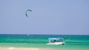 A glass bottom boat on the water while a kite surfer surfs by, the sky is blue, the water is calm.
