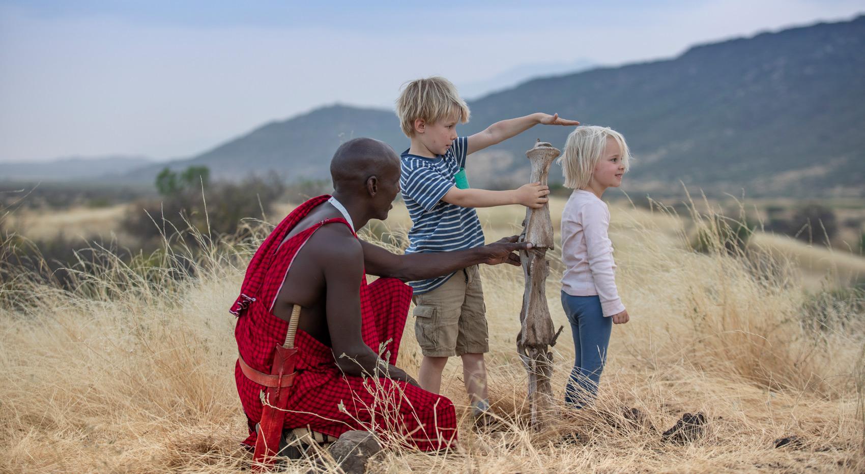 Children and a Masai man measuring the youngest child's hight with a large dry piece of wood.